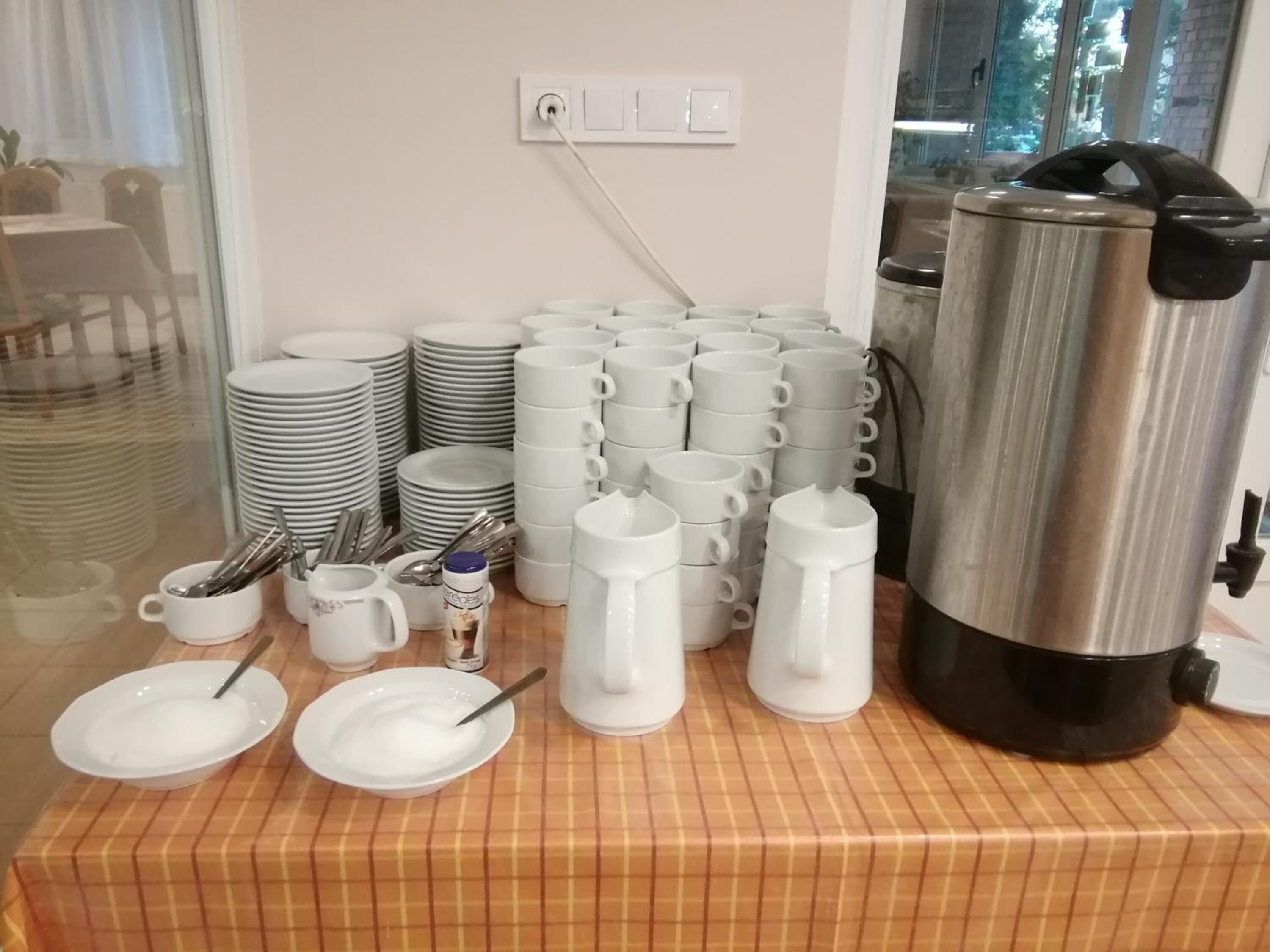 A buffet setup with stacks of white plates, bowls, and mugs, a coffee pot, and a can of creamer on a checkered tablecloth.