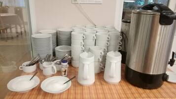 A buffet setup with stacks of white plates, bowls, and mugs, a coffee pot, and a can of creamer on a checkered tablecloth.