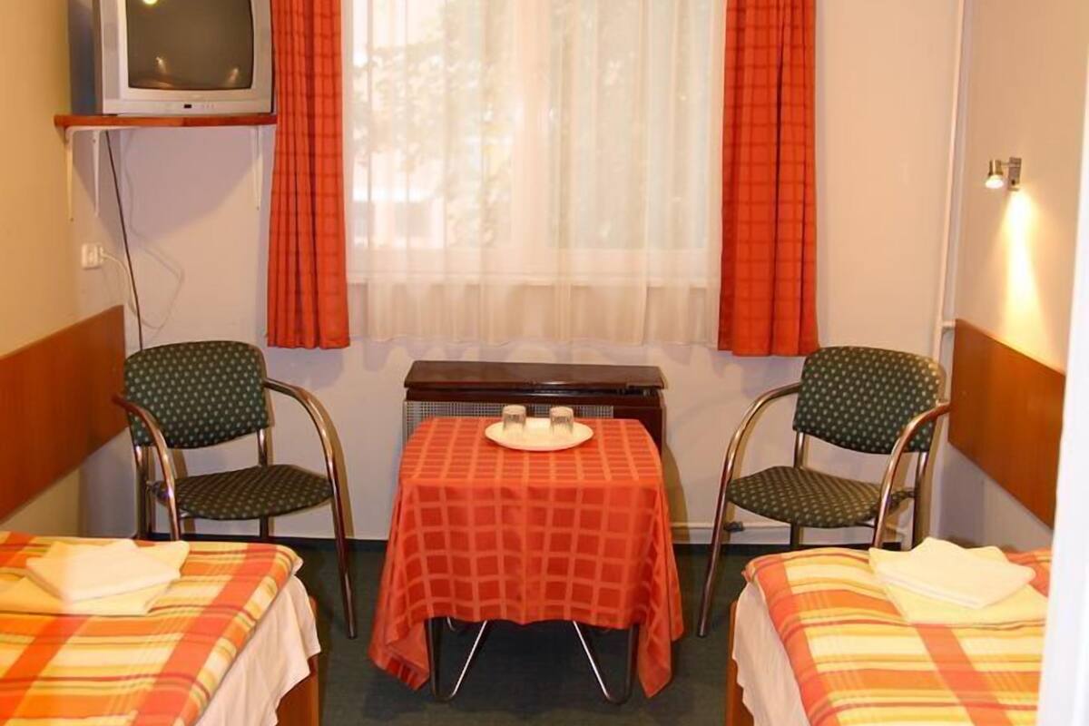 A small hotel room with two single beds, a small table with a red tablecloth, two chairs, a television on a shelf, and a window with curtains.