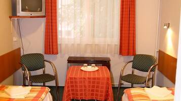 Une petite chambre d’hôtel avec deux lits, une table, des chaises, une télévision et une fenêtre avec des rideaux.