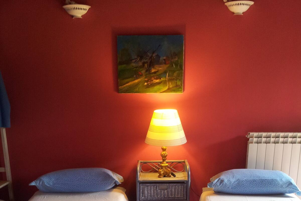 A room with two beds, each with a blue pillow and a white blanket. There is a small table with a lamp between the beds, and a painting on the wall above the table.