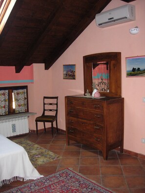 A bedroom with a wooden dresser, a chair, a radiator, a window with floral curtains, and a wall-mounted air conditioner. - Il Pioppo e la Fonte (Castello d'Agogna)