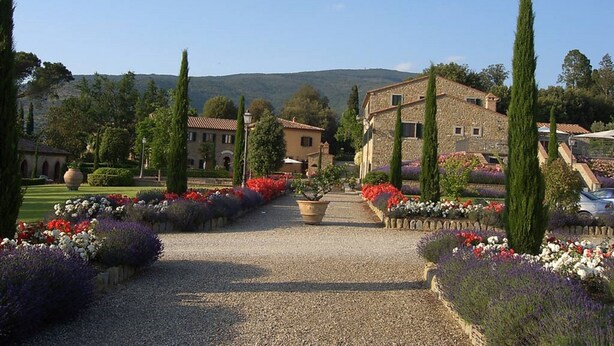 Un jardin avec un chemin de gravier menant à un bâtiment en pierre, entouré de grands cyprès et de parterres de fleurs aux couleurs vives.