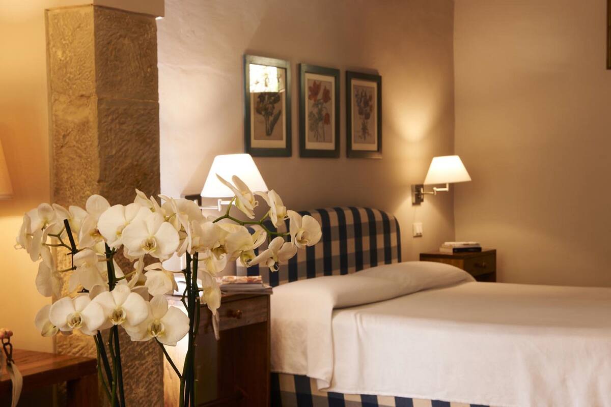 Een hotelkamer met een bed, twee bedlampen, een nachtkastje met lamp, een vaas met witte bloemen en lijstjes met afbeeldingen aan de muur.