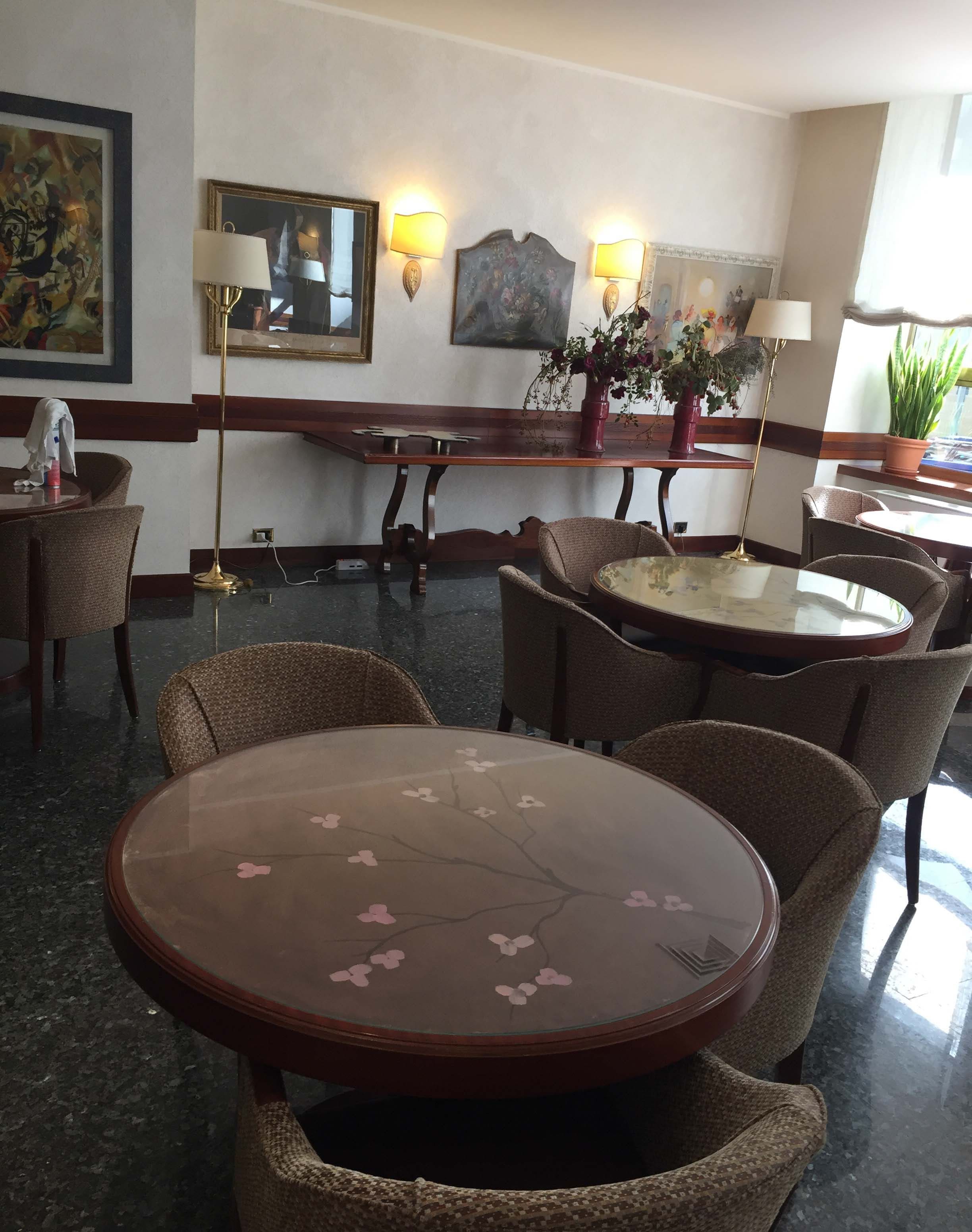 Un hall d'hôtel avec des tables rondes, des chaises en osier, des lampes fixées au mur et un grand tableau.