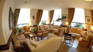 A spacious living room with multiple sofas and armchairs, large windows with curtains, and several potted plants. - Hotel Bristol (Milan)