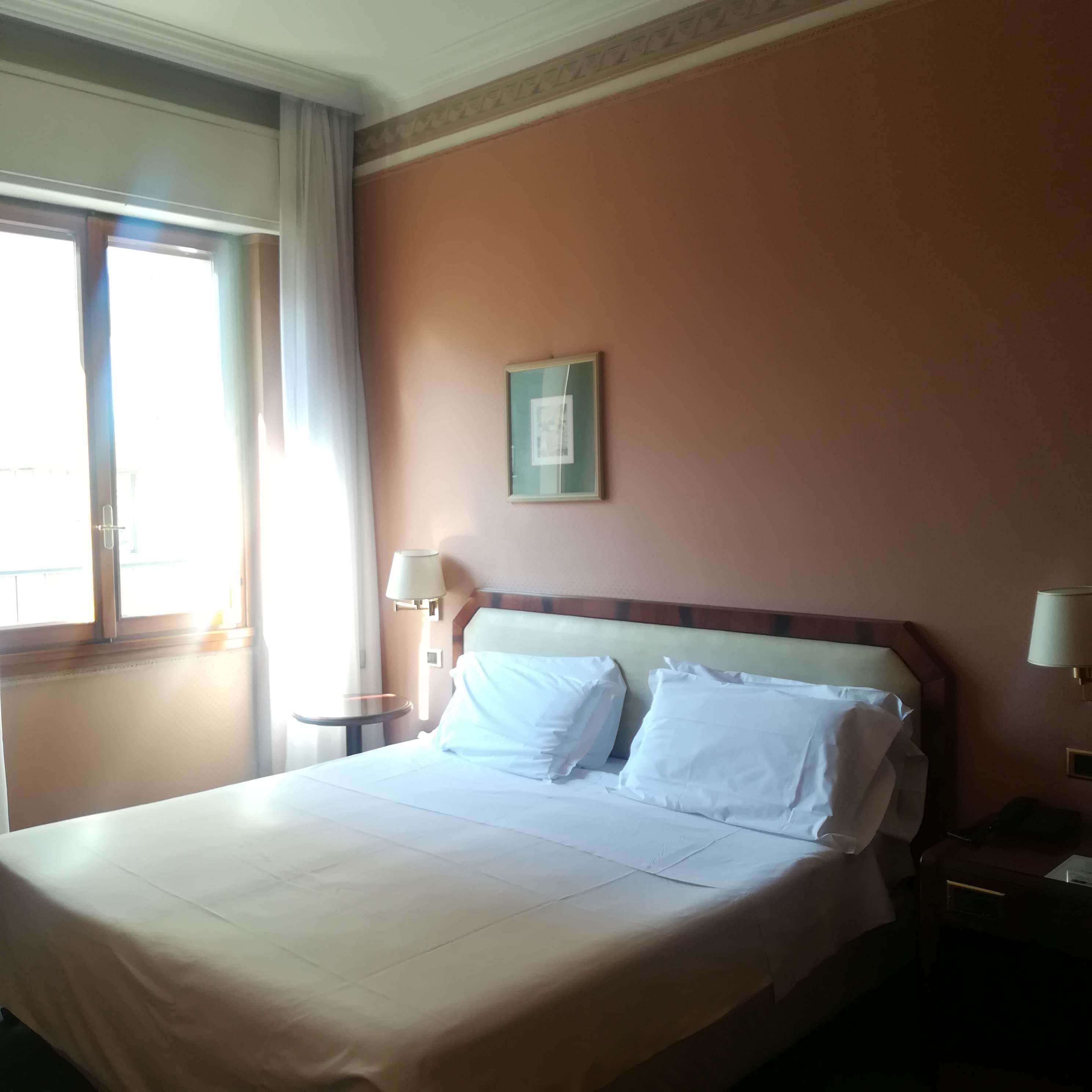 A hotel room with a bed, two bedside lamps, a window with curtains, and a framed picture on the wall.