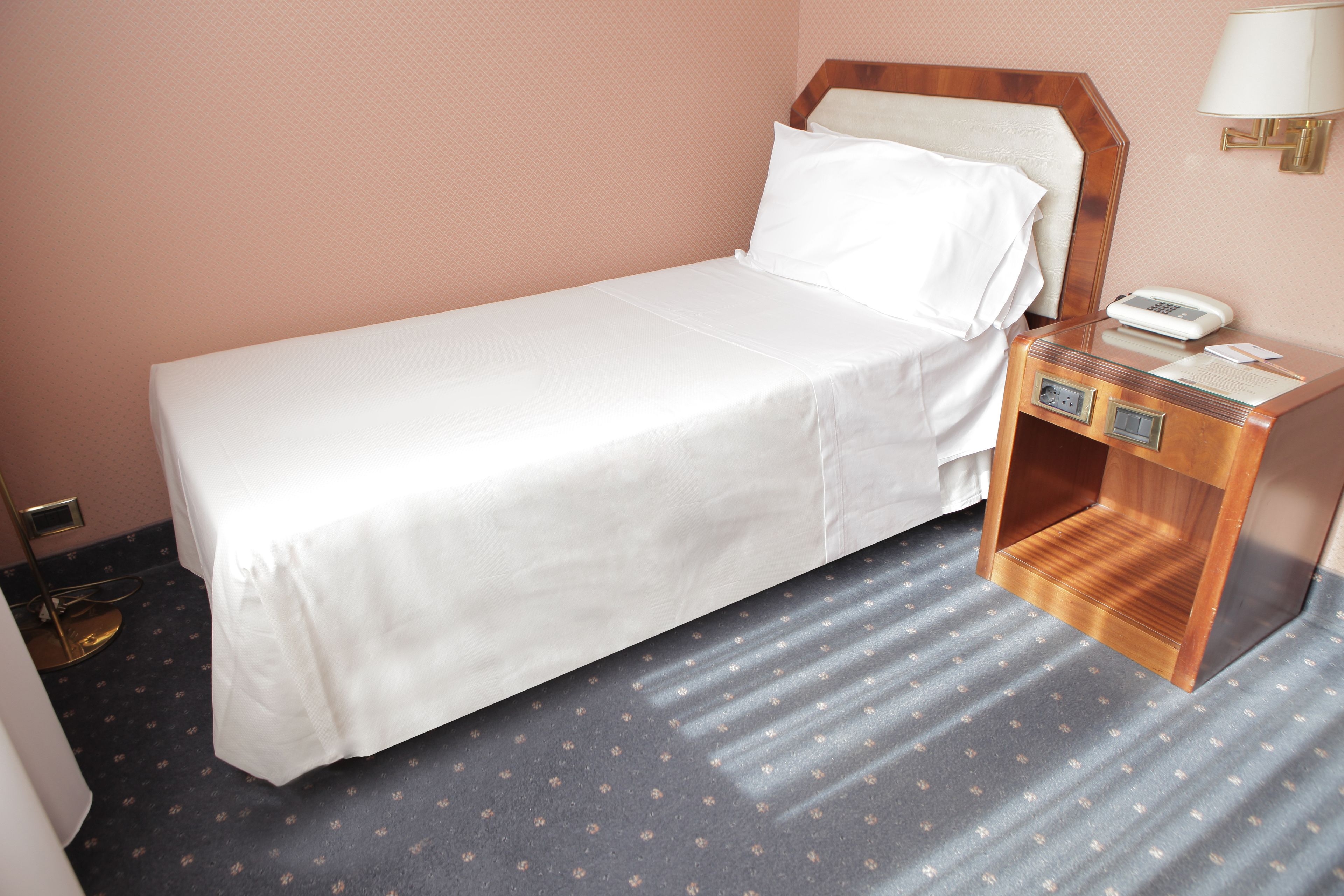 A single bed with white linens, a bedside table with a lamp, and a small drawer unit.