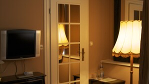 Una habitación de hotel con televisión sobre un mueble bajo, una puerta con espejo y una mesita de noche con lámpara.