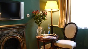 Una habitación con una televisión montada en la pared, una silla de madera con cojín, una mesa con lámpara, un jarrón con flores y una chimenea.