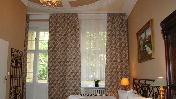 Una habitación de hotel con cama, ventana con cortinas, radiador, lámpara y un cuadro en la pared.