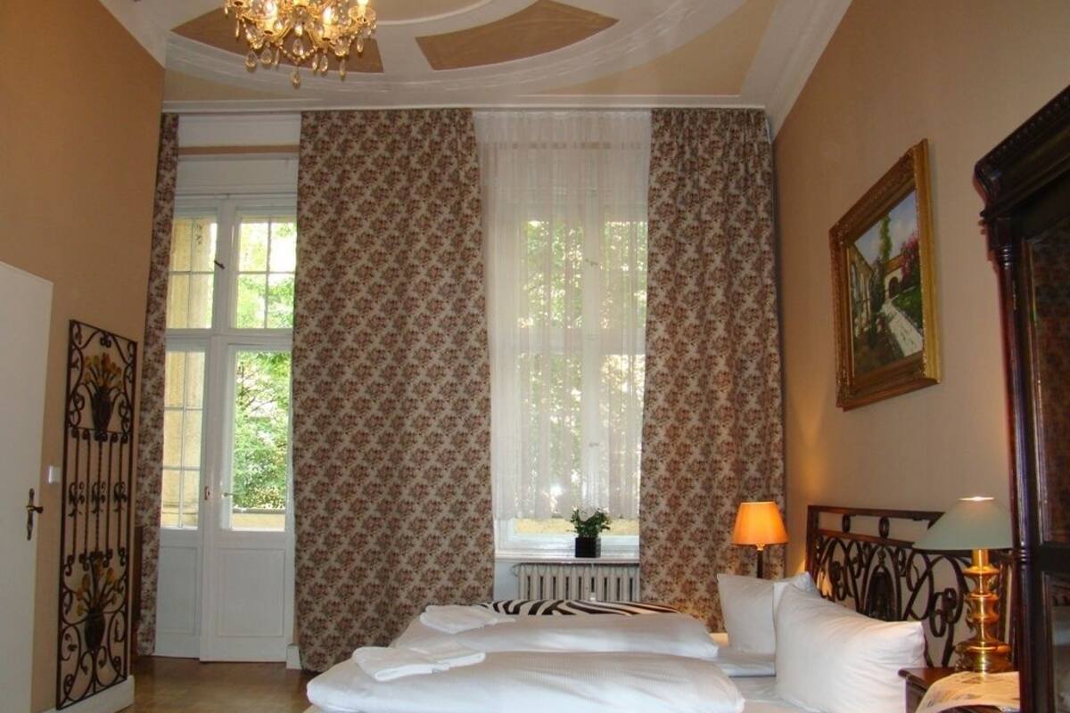 Une chambre d’hôtel avec un lit, une fenêtre avec des rideaux, un radiateur, une lampe et un tableau au mur.