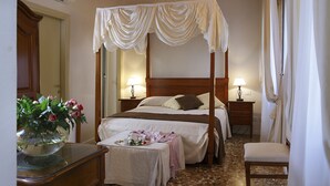 Una cama con dosel, mesitas de noche con lámparas, una pequeña mesa con un jarrón de flores y una silla.