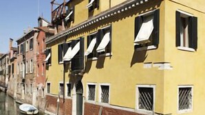 Un edificio junto a un canal, con fachada amarilla, varias ventanas y el agua del canal a la vista.