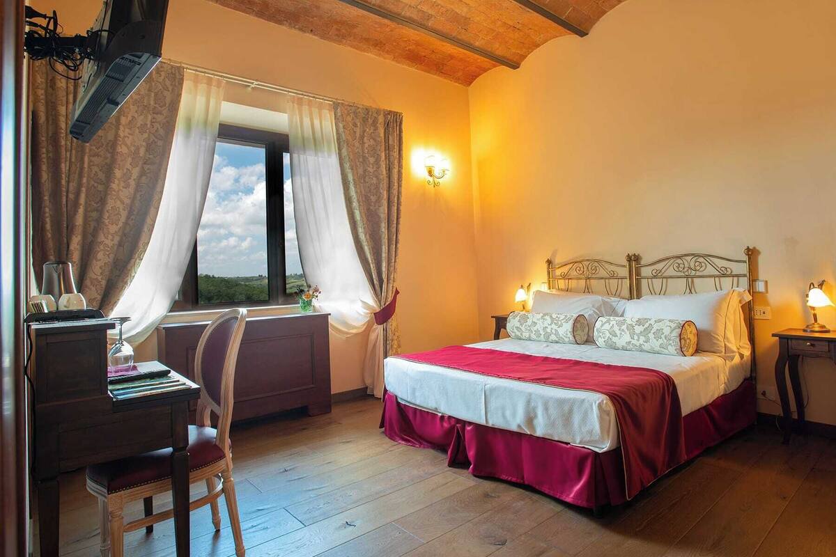 A bedroom with a wooden bed, a desk with a chair, a window with curtains, and a wall-mounted lamp.