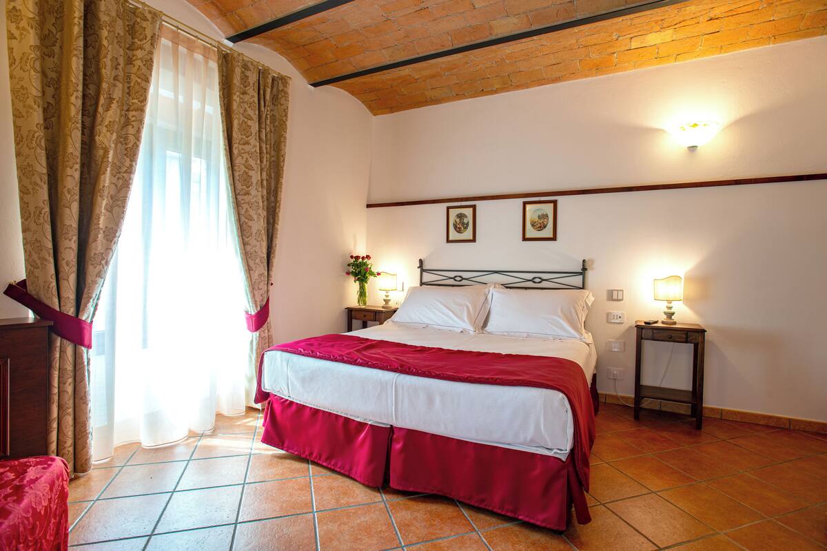 A bedroom with a bed, curtains, a nightstand, a lamp, and a wall-mounted light fixture.