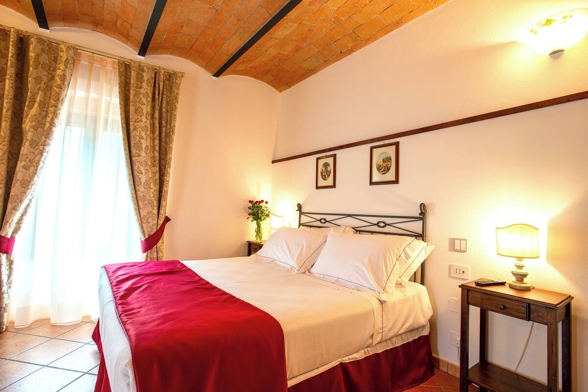 A neatly made bed with a red bedspread, a bedside table with a lamp, and two framed pictures on the wall.