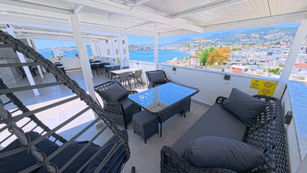 A covered outdoor seating area with wicker furniture, a glass-top table, and a view of the sea and cityscape. - Merhaba Pansiyon (Bodrum)