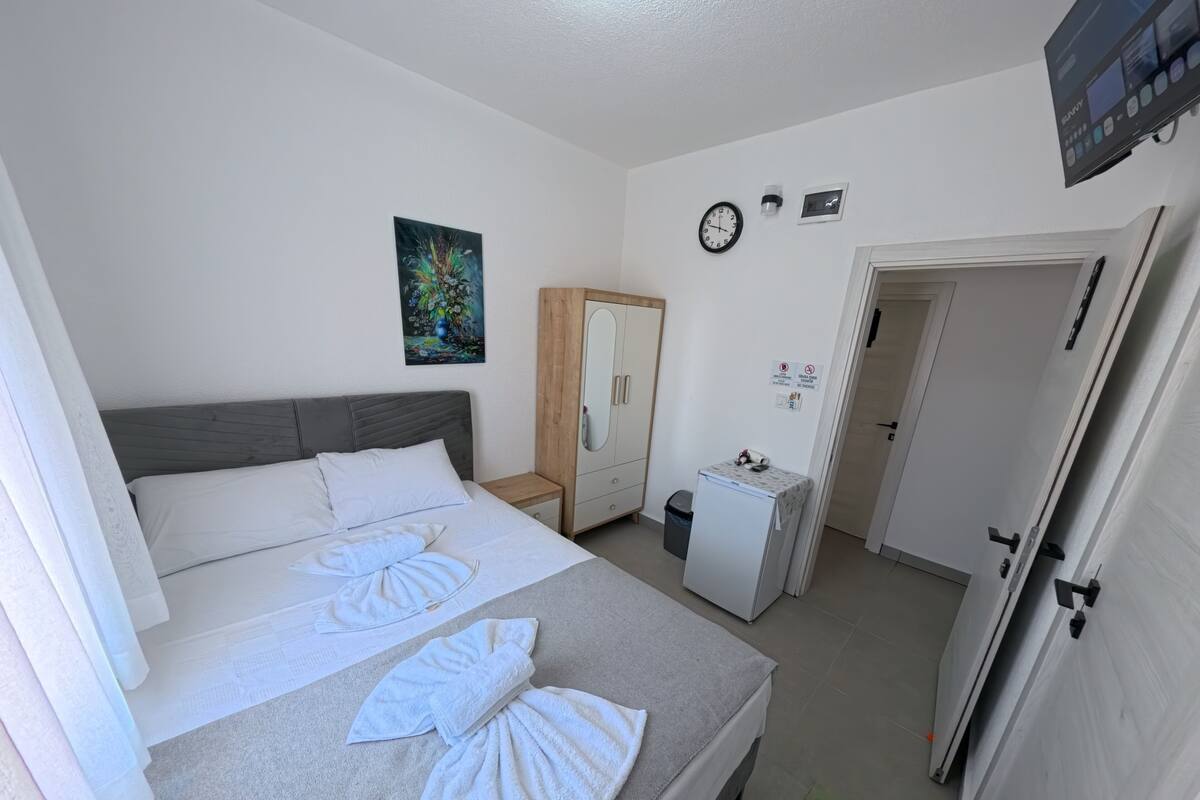 A hotel room with a bed, a small desk, a wardrobe, a clock, and a painting on the wall.
