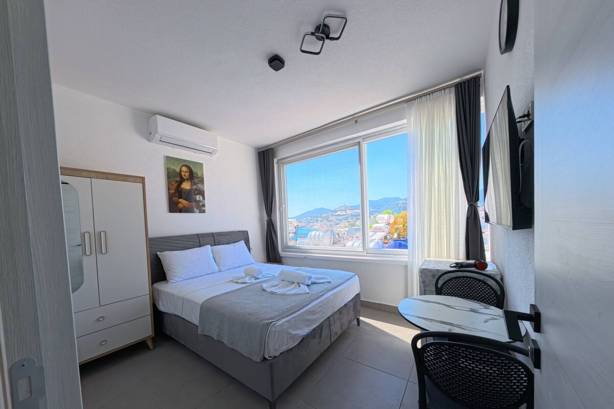 A hotel room with a bed, a desk, a chair, a wardrobe, a painting, a window with a city view, and an air conditioning unit.