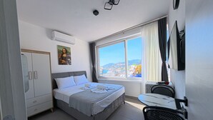 A hotel room with a bed, a desk, a chair, a wardrobe, a painting, a window with a city view, and an air conditioning unit. - Merhaba Pansiyon (Bodrum)
