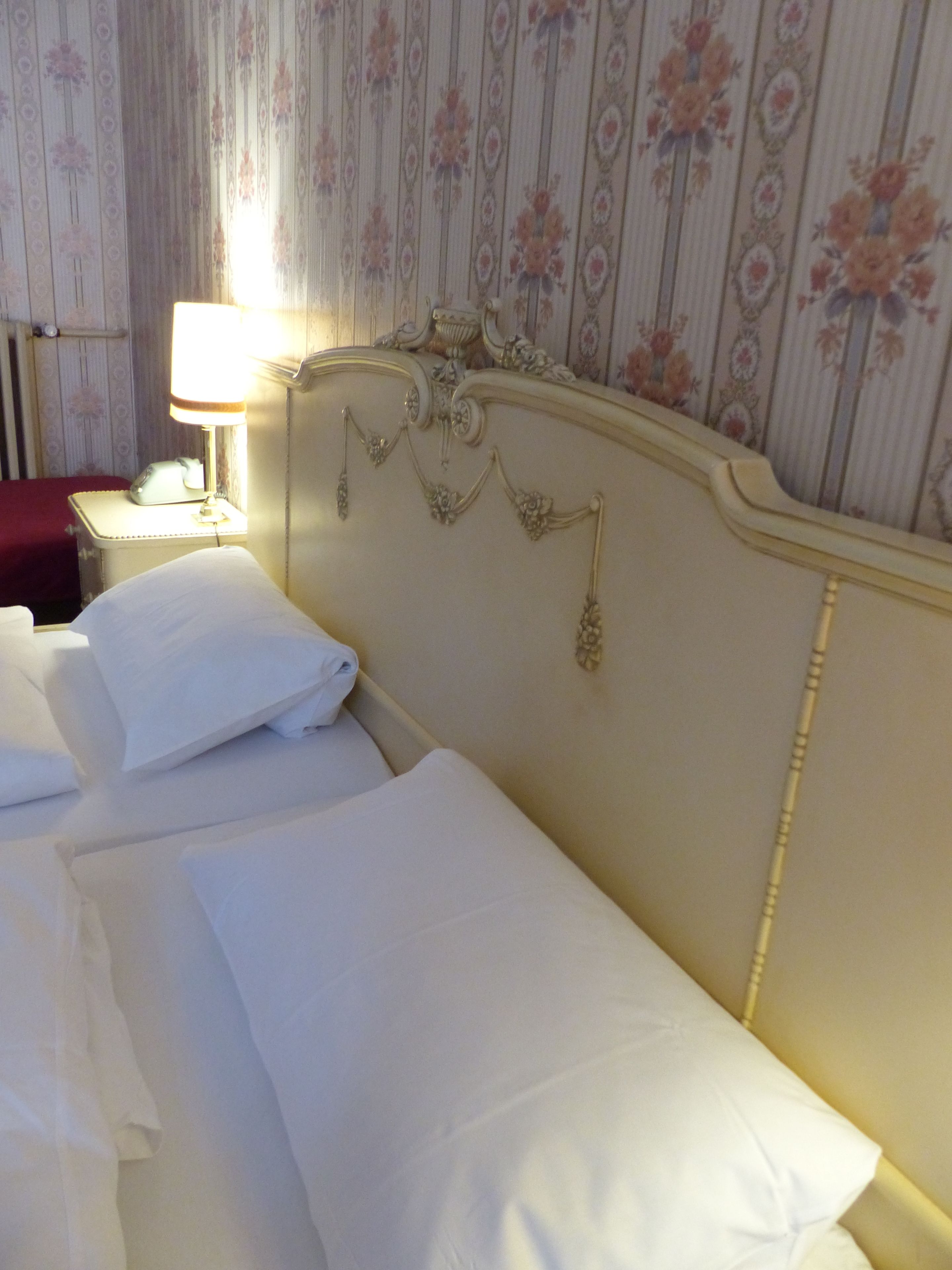 Una habitación de hotel con dos camas ordenadas, una lámpara de noche y papel tapiz con motivos florales.