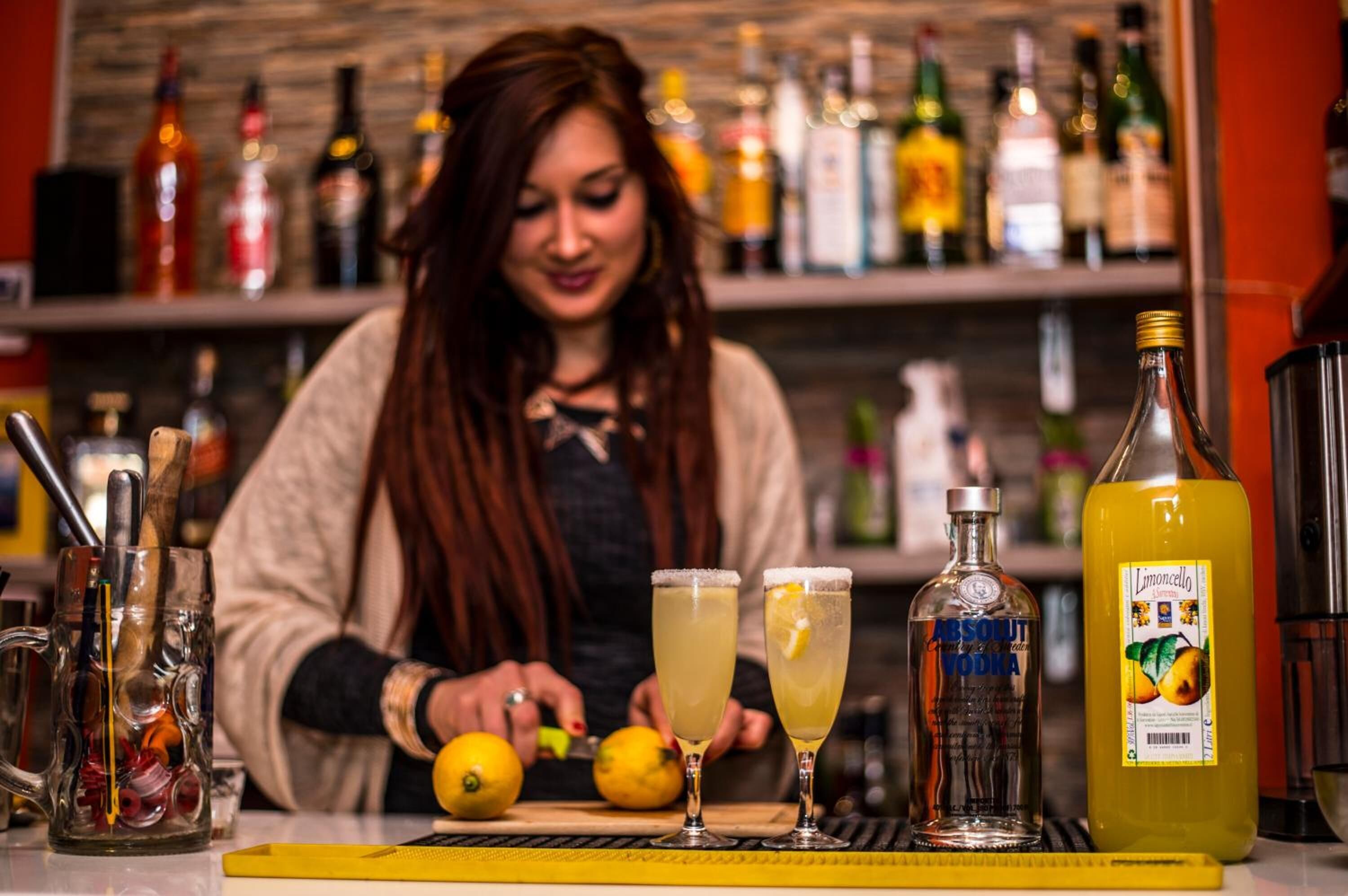 Un bar con un cantinero preparando bebidas, una variedad de botellas y una botella grande de jugo de limón sobre la barra.