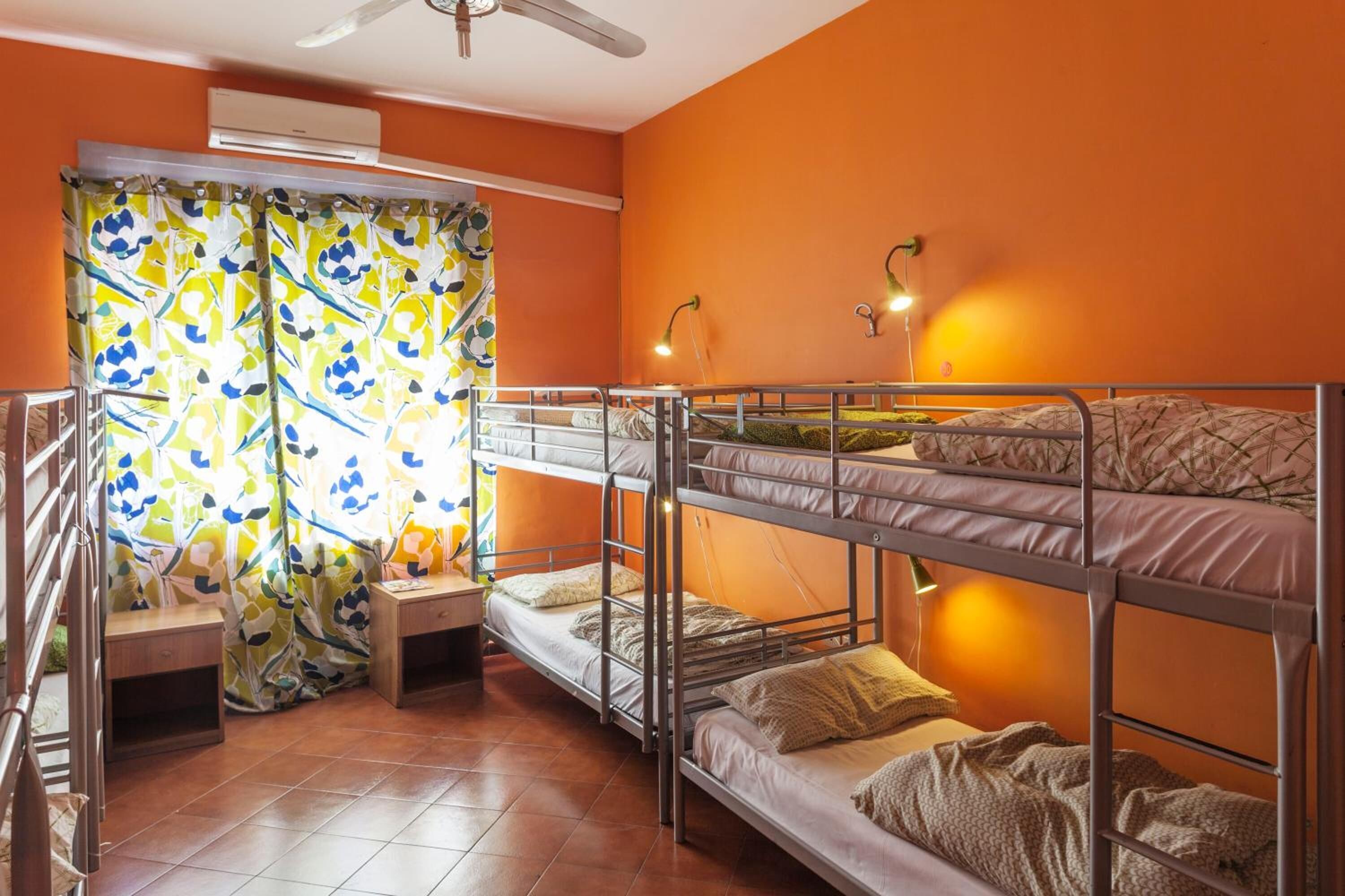 A dormitory room with bunk beds, a window with curtains, a small table, and a ceiling fan.