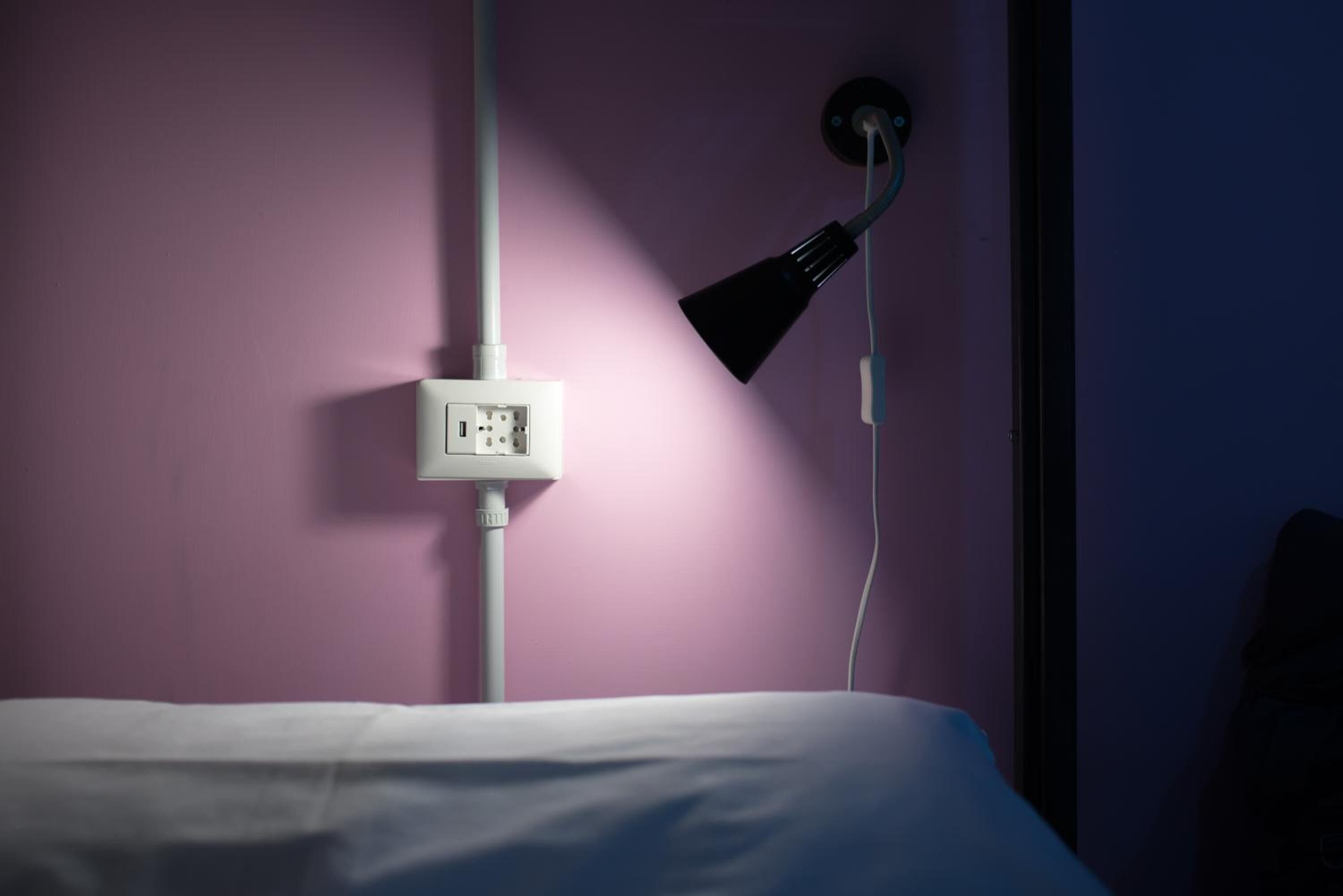 A bed with a white sheet, a wall socket, and a black lamp on a purple wall.
