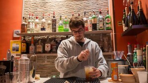Eine Bar mit Regalen voller Spirituosenflaschen, ein Barkeeper, der einen Cocktail zubereitet, und ein Menüschild im Hintergrund.