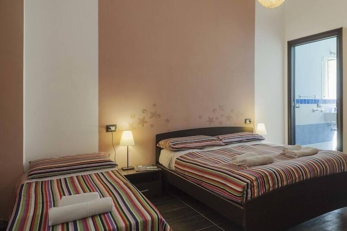 Ett hotellrum med två sängar, båda med randiga sängkläder och en hoprullad handduk. Det finns väggmonterade lampor och ett stort fönster med utsikt över omgivningen.