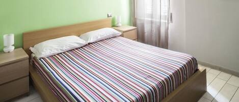 A single bed with a colorful striped bedspread, a wooden headboard, and a nightstand with a white lamp.