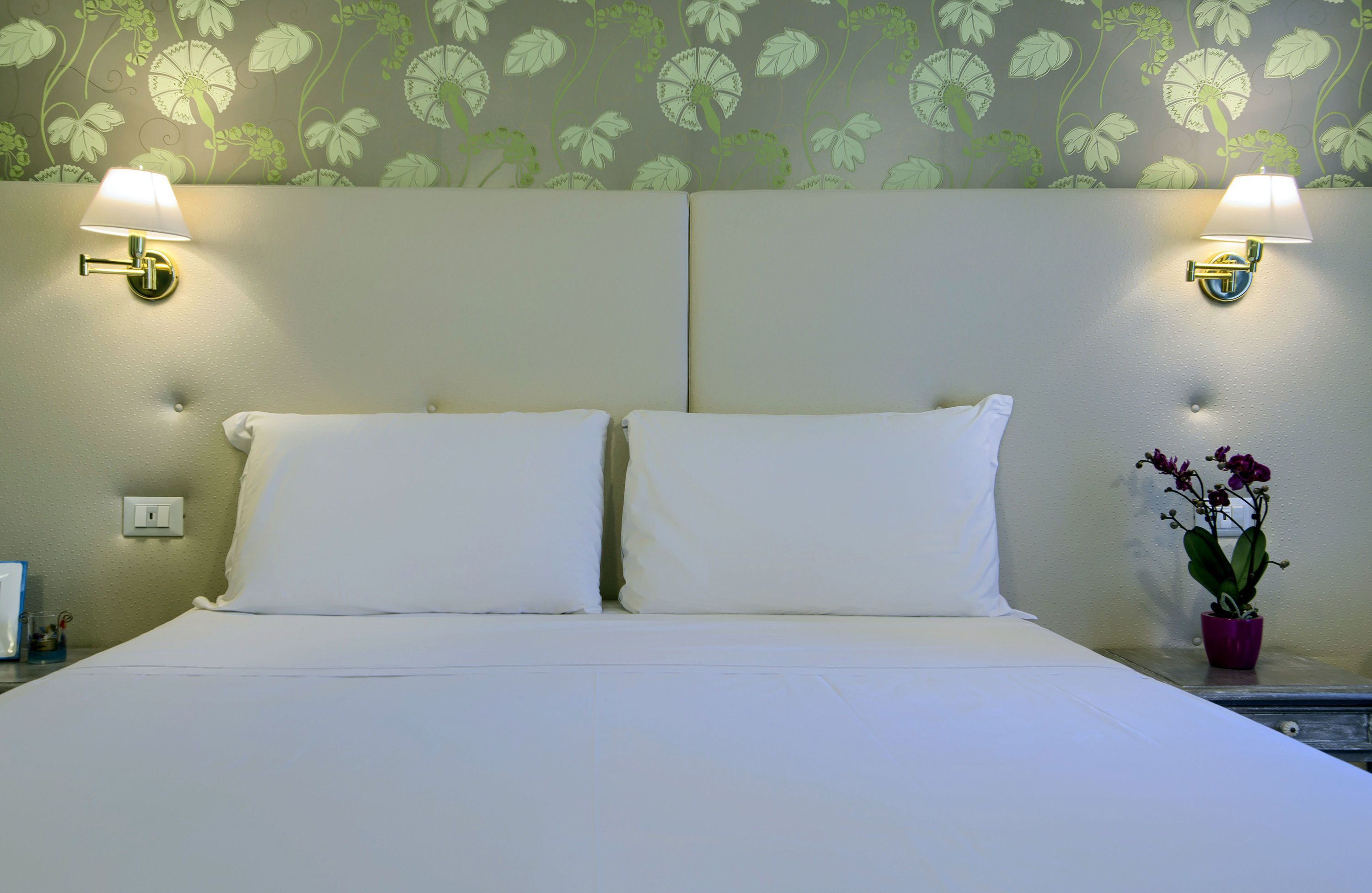 Hotellihuone, jossa on suuri sänky, kaksi seinälle asennettua lamppua ja ruukkuorkidea yöpöydällä.