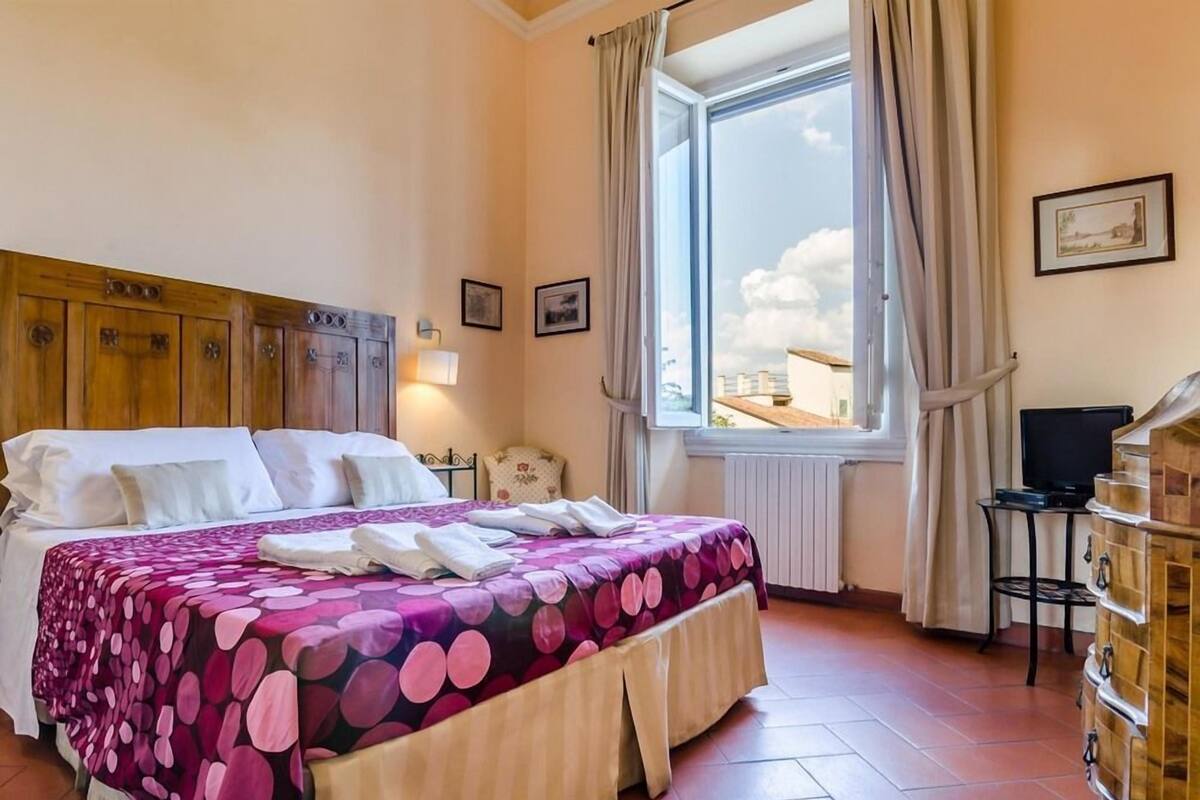 A hotel room with a bed, a window with curtains, a desk with a computer, and a wooden cabinet.