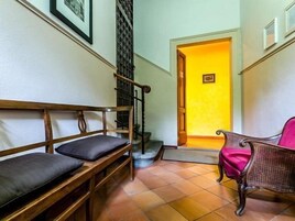 Un pasillo con un banco de madera y una silla de mimbre, suelo de baldosas y una puerta que da a una habitación con pared amarilla.