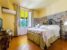 Un dormitorio con cama doble, una mesita de noche con lámpara, ventana con cortinas, pared de piedra y cómoda de madera.