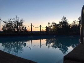 Una zona piscina all'aperto tranquilla con una vista libera sul tramonto, circondata da alberi e una recinzione.