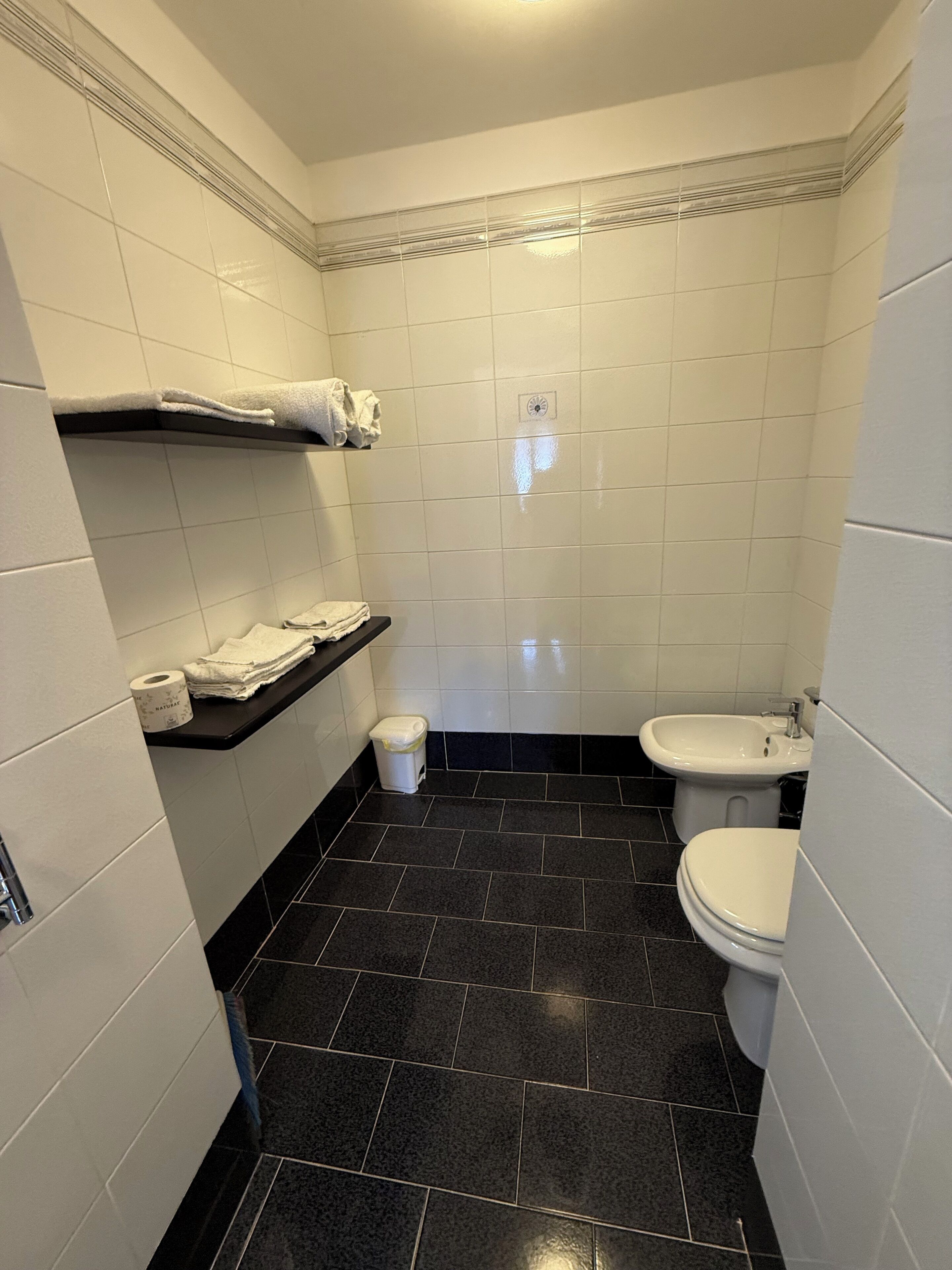 Ein kleines Badezimmer mit weißen Fliesenwänden, einem schwarzen Fliesenboden, einer Toilette, einem Bidet, einem Ablagefach mit Handtüchern und einem Mülleimer.