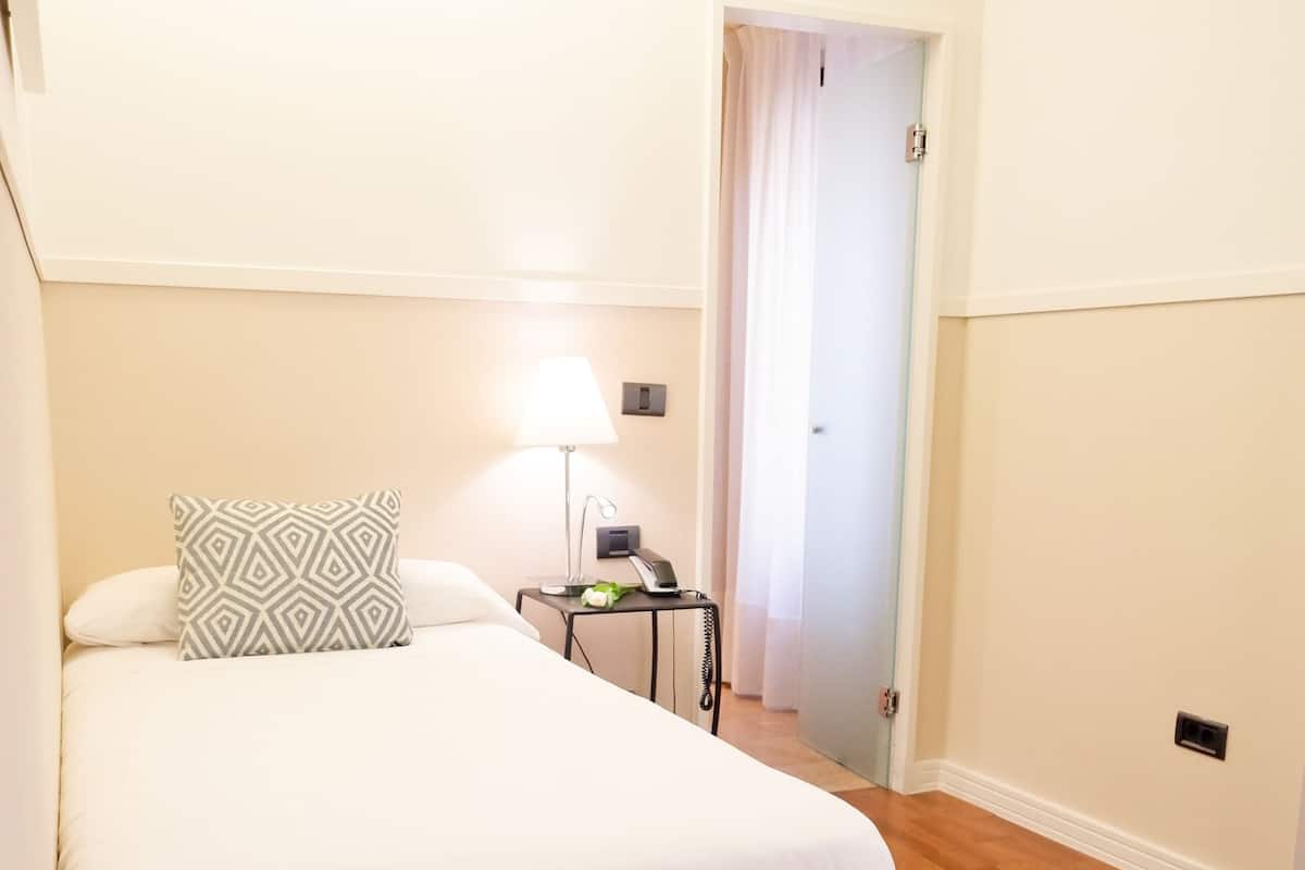 A bedroom with a bed, a bedside table with a lamp, and a door leading to another room.