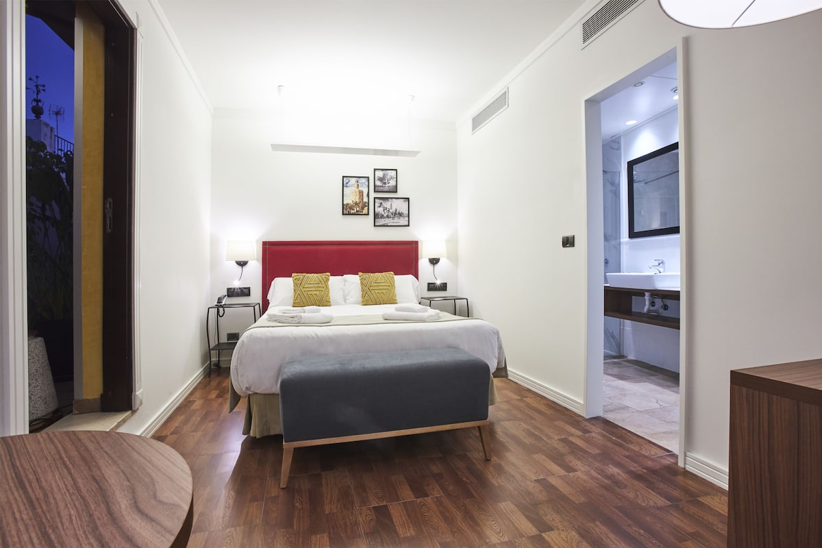 A modern hotel room with a wooden floor, a red headboard, a bed with white linens, a bedside table with a lamp, and a view of the outside.