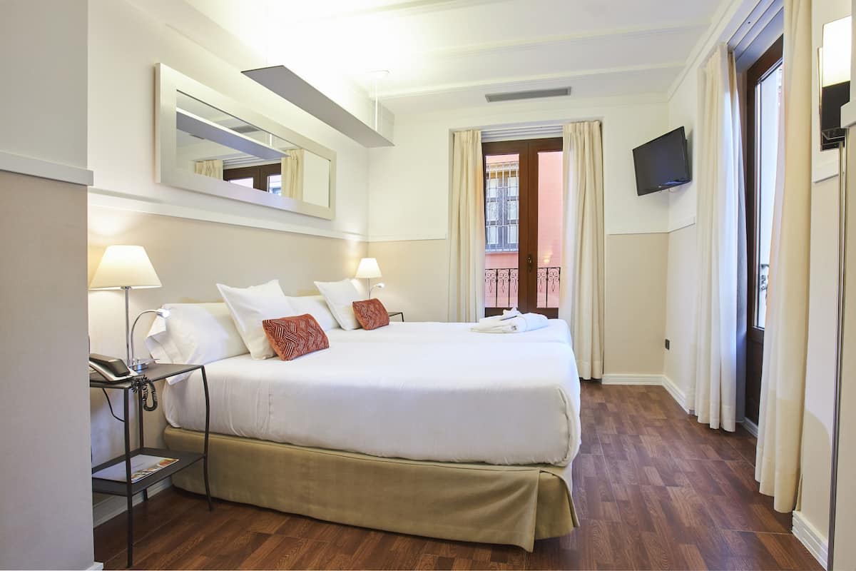 A hotel room with a bed, bedside lamps, a bedside table with a phone, a TV mounted on the wall, and a balcony with curtains.