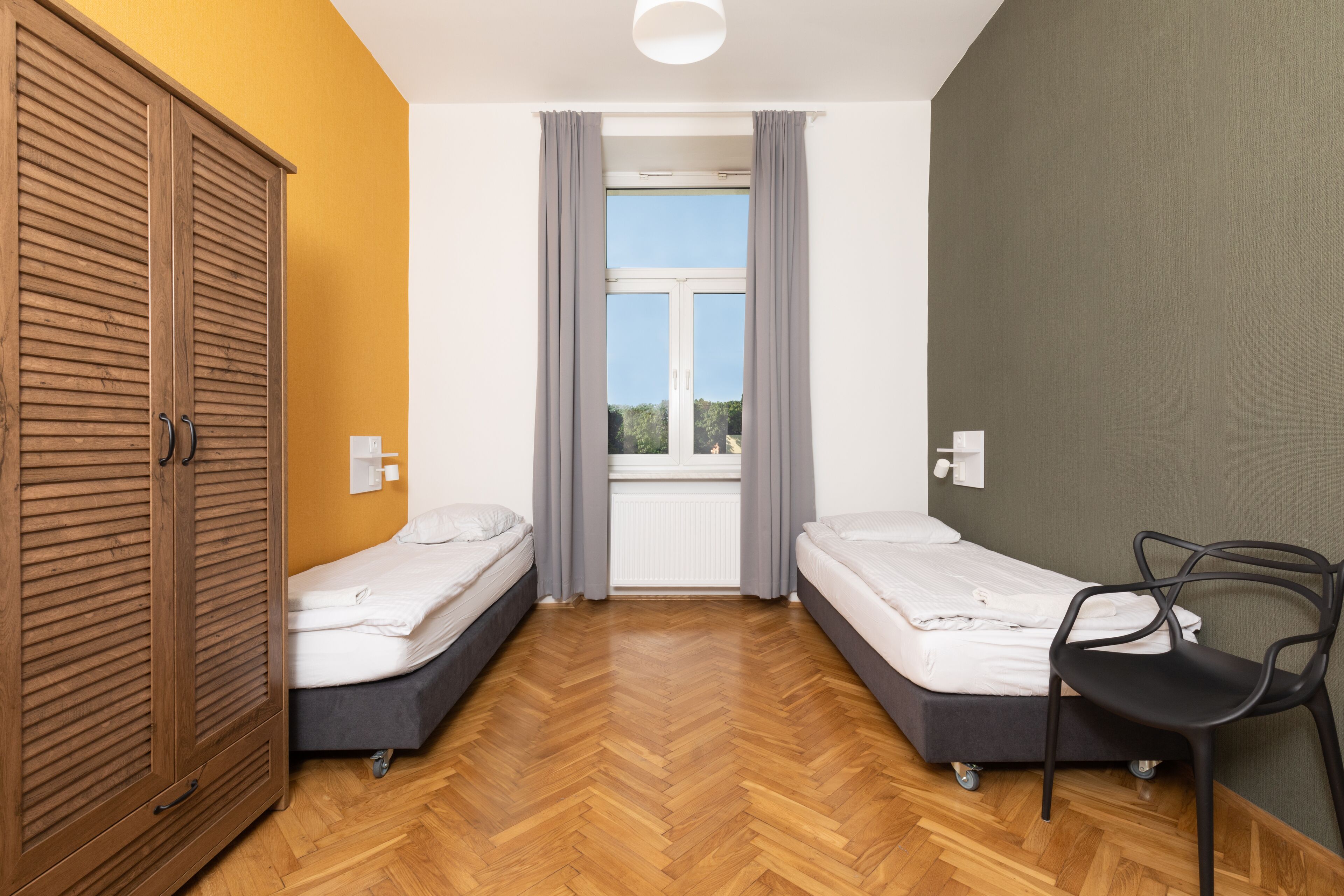 A room with two beds, a wooden wardrobe, a chair, and a window with curtains.