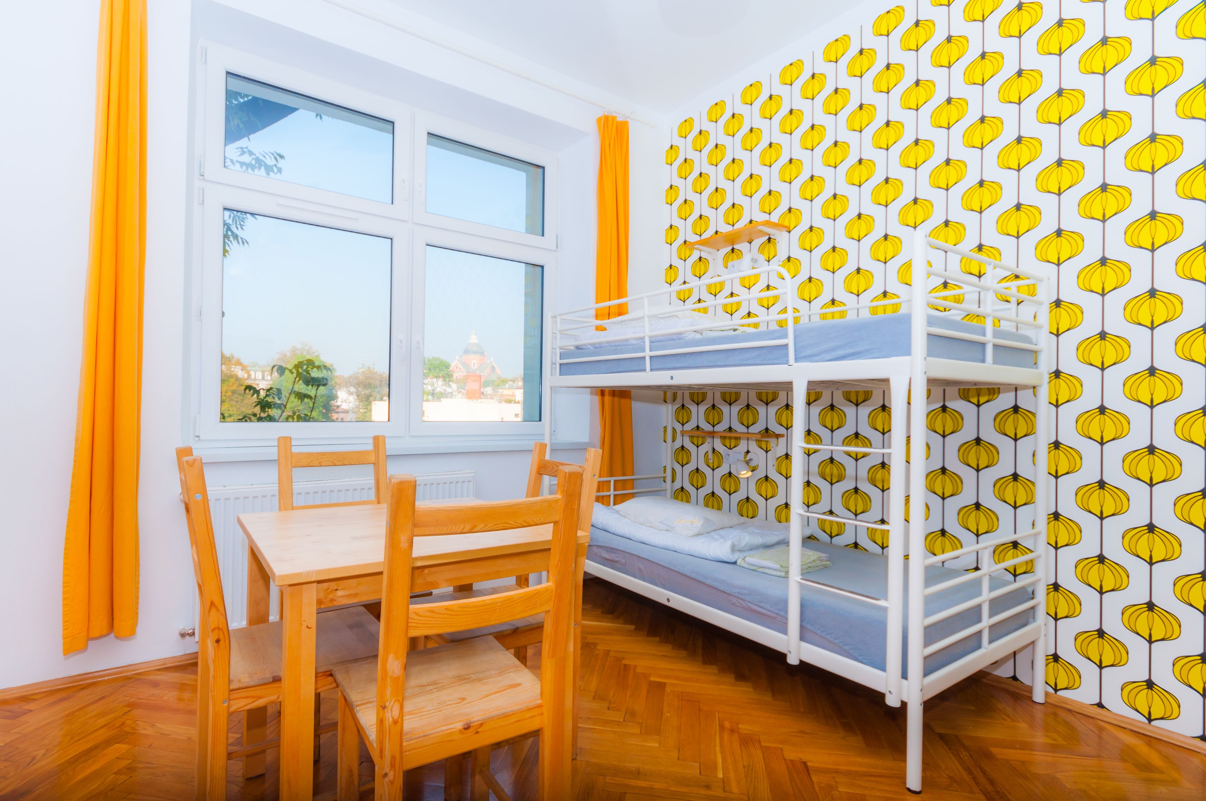 A room with a bunk bed, a wooden table, and chairs. There is a window with curtains and a view of buildings outside.