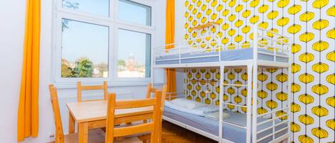 A room with a bunk bed, a wooden table, and chairs. There is a window with curtains and a view of buildings outside.