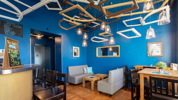 A restaurant interior with a blue wall, wooden ceiling, and hanging pendant lights. There are wooden tables and chairs, a counter with a chalkboard menu, and a sofa. - Hostel Atlantis (Kraków)