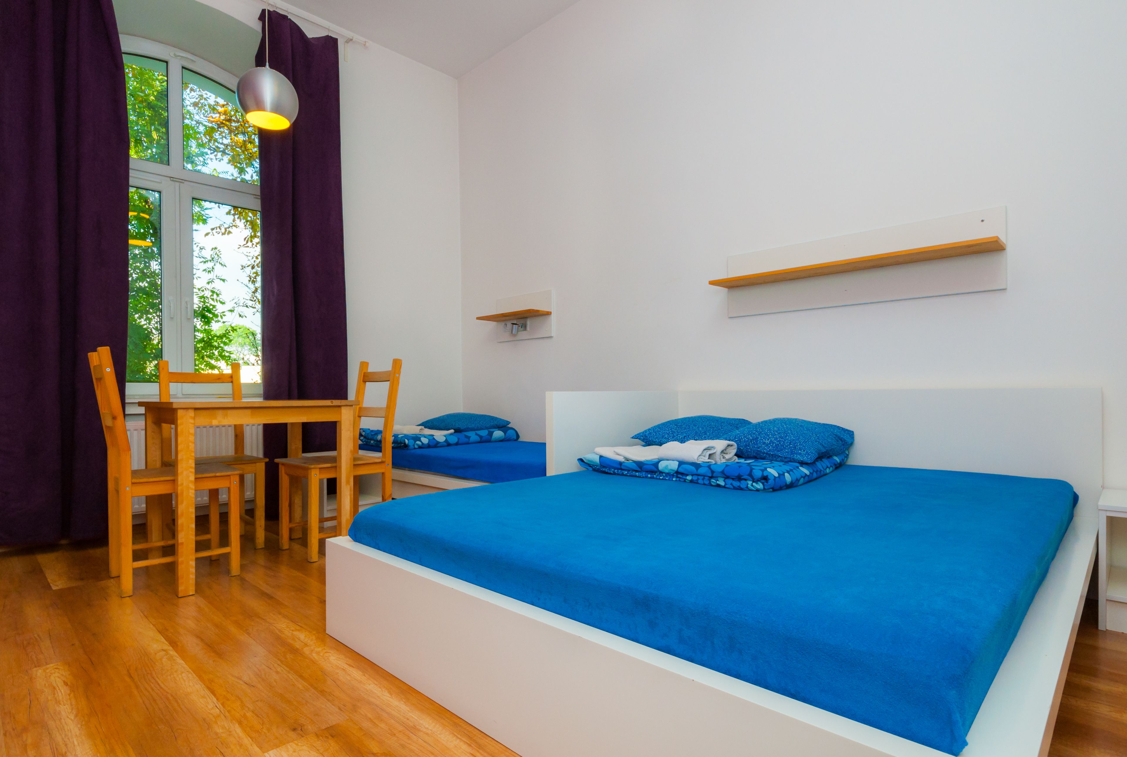 A bedroom with two beds, a wooden table, chairs, a window with curtains, and a hanging light fixture.