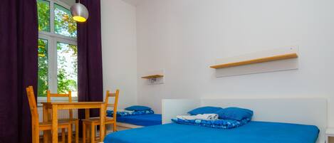 A bedroom with two beds, a wooden table, chairs, a window with curtains, and a hanging light fixture.
