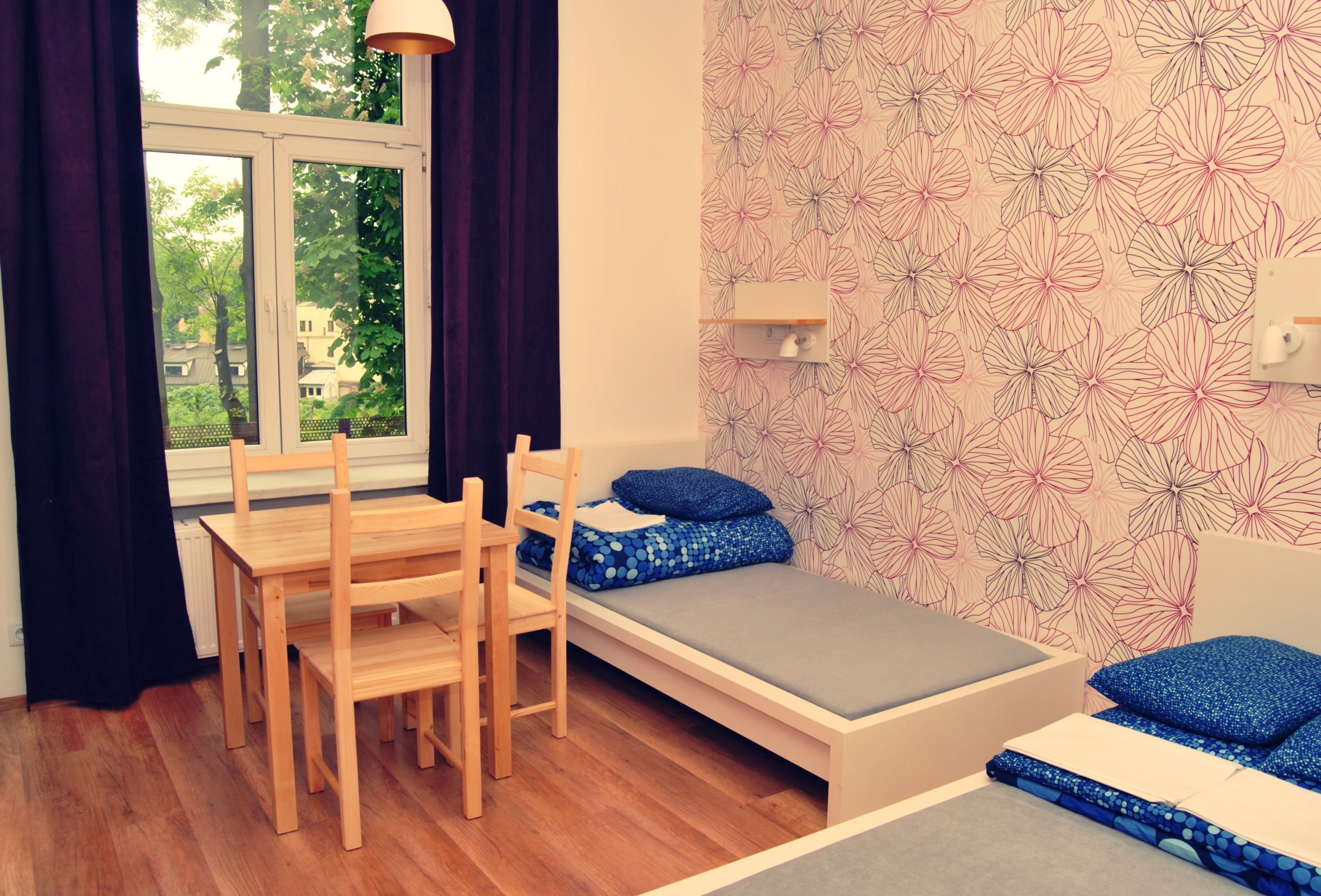 Una habitación pequeña con una cama individual, una silla de madera, una mesa y un ventanal con cortinas.