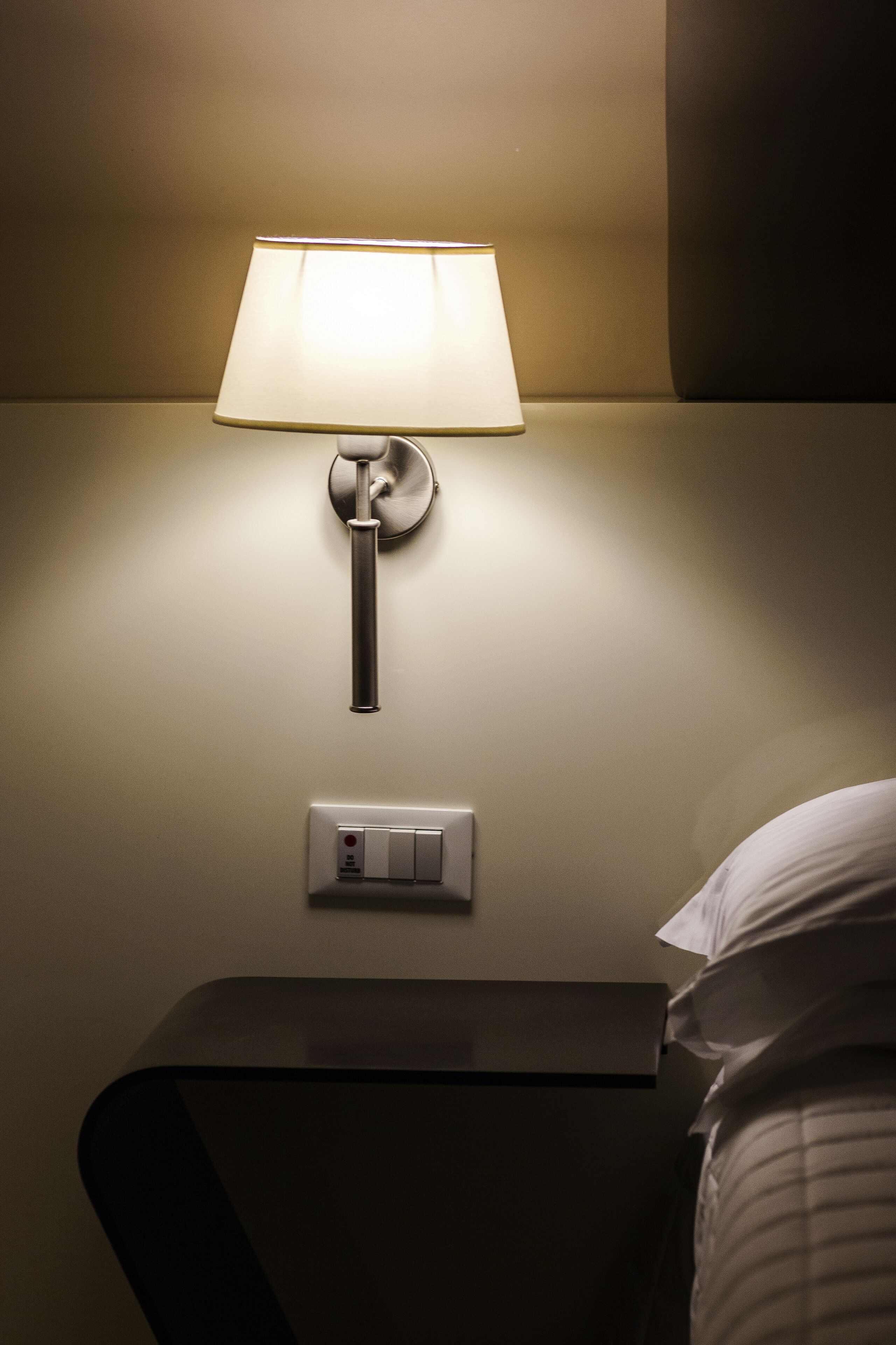 A wall-mounted lamp with a switch below it, a bedside table, and a partially visible bed.
