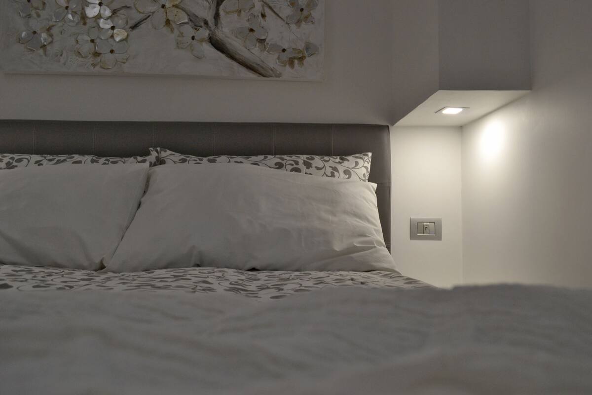 A bedroom with a bed, pillows, a wall-mounted artwork, and a light switch.